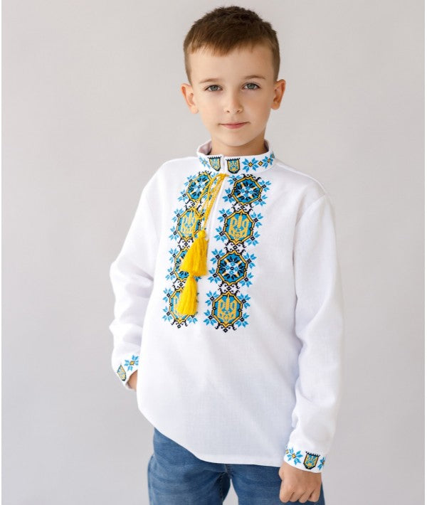 A boy wearing a white embroidered shirt with the Ukrainian coat of arms, featuring blue and yellow embroidery, a yellow tassel on the collar, and the shirt is complemented by decorative tassels on the collar and cuffs.
