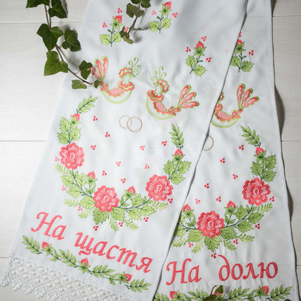 A traditional embroidered Ukrainian wedding towel with floral patterns and wedding ring motifs in green and gold threads, displayed on a white background with green foliage accents.