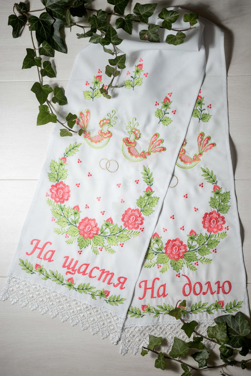 A traditional embroidered Ukrainian wedding towel with floral patterns and wedding ring motifs in green and gold threads, displayed on a white background with green foliage accents.