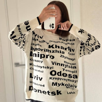 Mirror selfie of a woman in a cream Ukrainian cities oversized sweater with black lettering