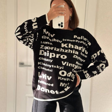 Mirror selfie close-up of a woman in a black Ukrainian cities knit sweater with white lettering
